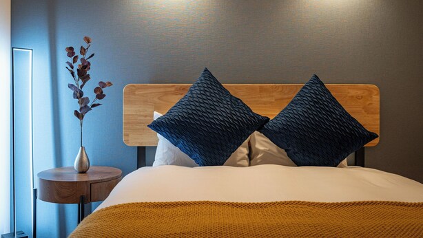 Una cama bien hecha con cabecera de madera, dos almohadas azul oscuro y una mesita redonda con un florero de hojas al lado.