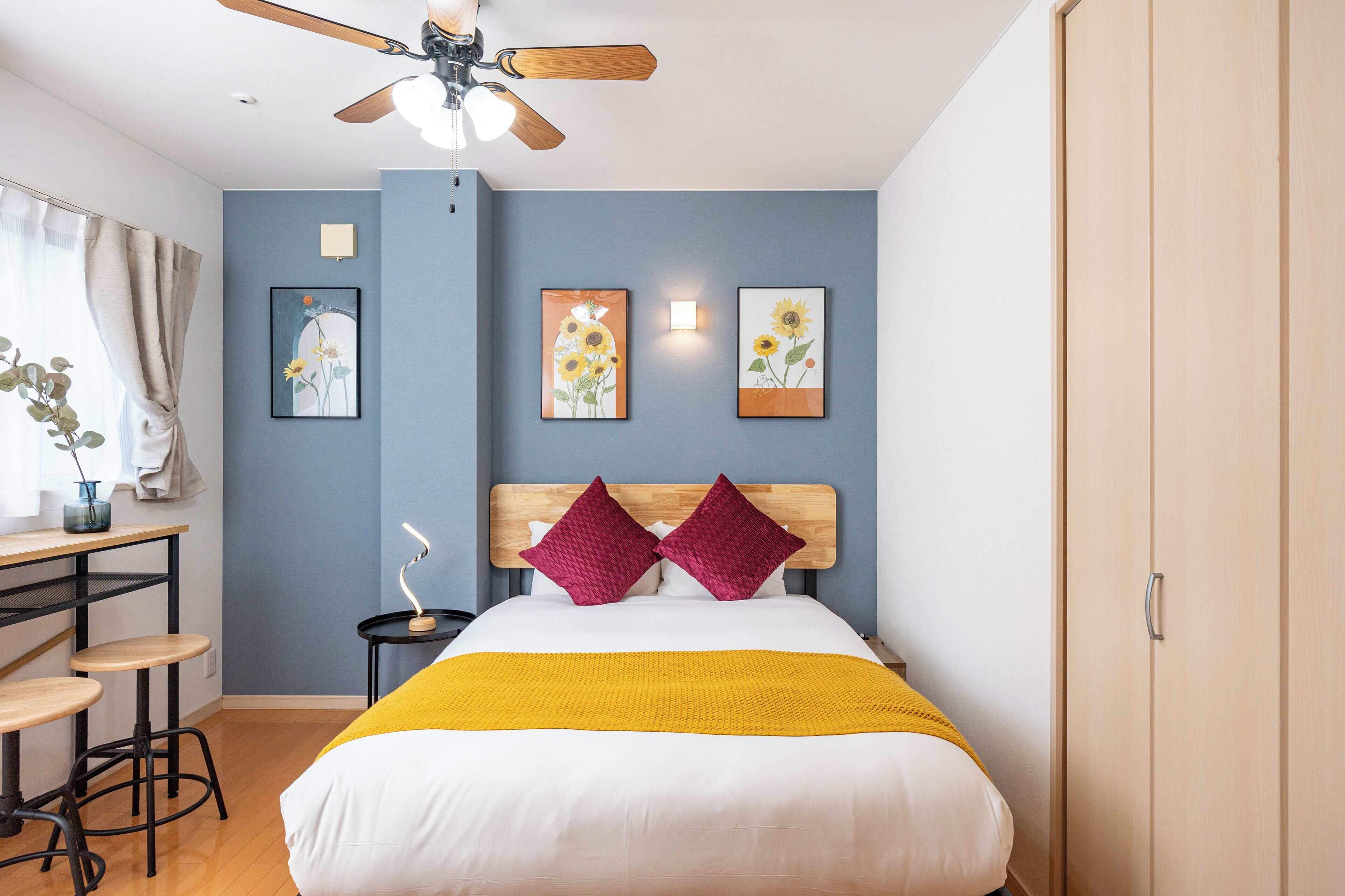 A bedroom with a bed, a desk with stools, a ceiling fan, and artwork on the walls.