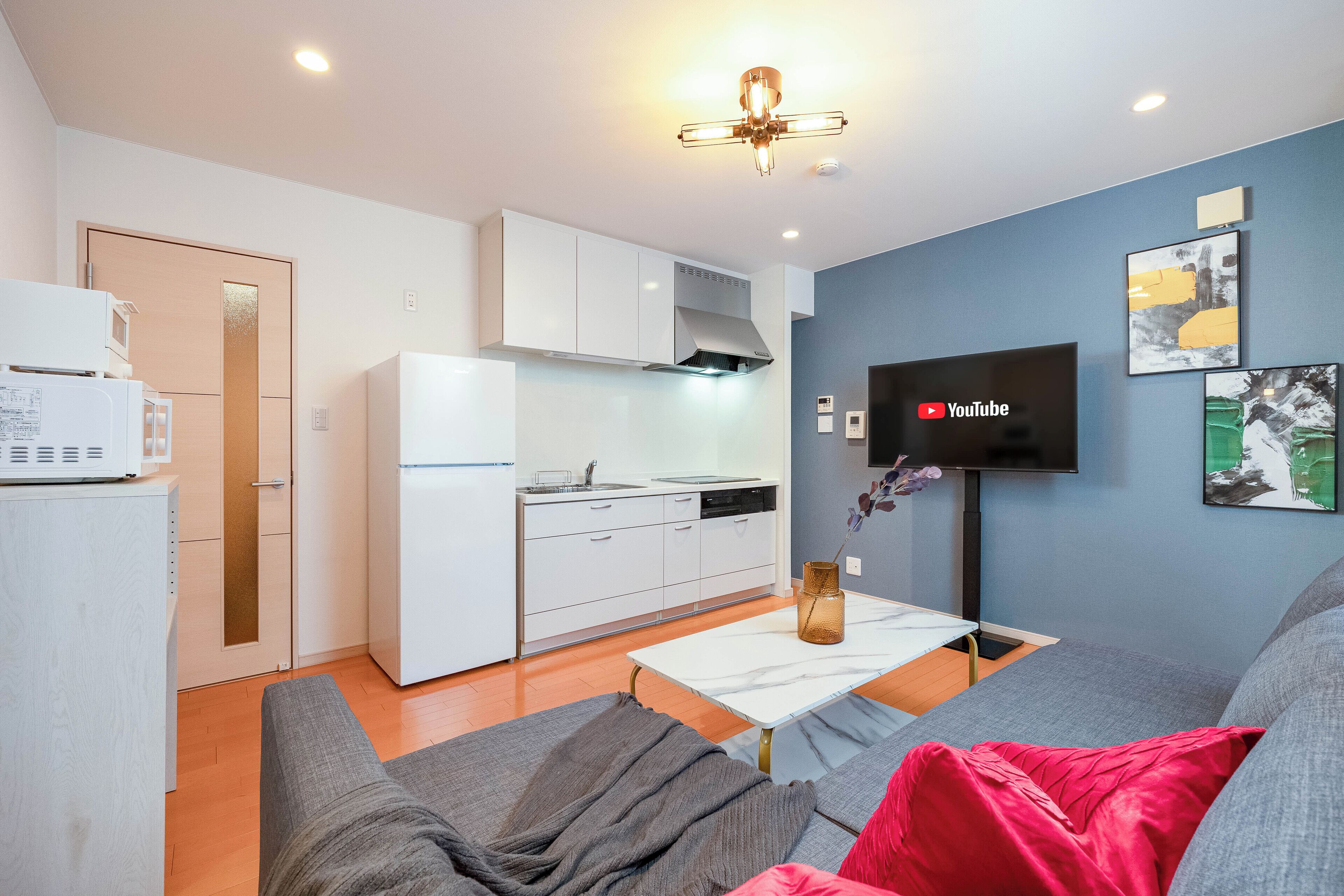 A modern kitchen with white appliances, a dining area with a white table and grey sofa, and a flat-screen TV mounted on the wall.