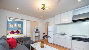 Una habitación moderna y bien iluminada con un sofá gris, una mesa de centro blanca y una zona de cocina con armarios y electrodomésticos blancos.