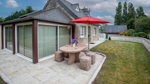 A stone patio with a round table and four stools, a red umbrella, and a house with a sloped roof and large windows.