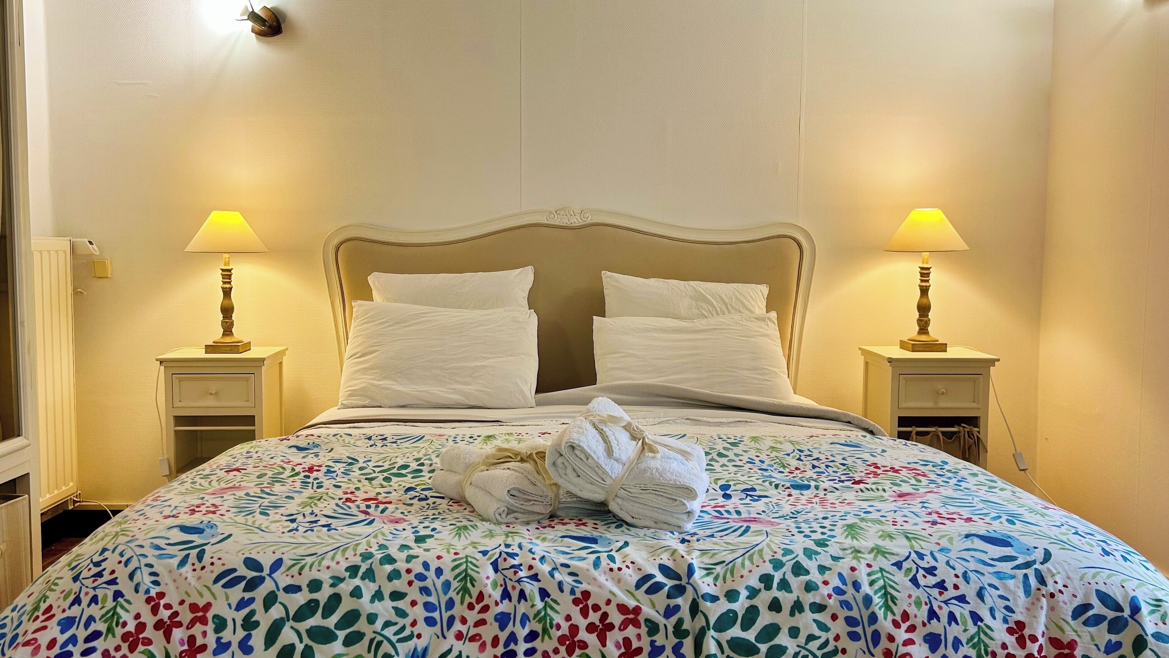 Una cama bien hecha con una colcha floral, dos almohadas blancas y una toalla doblada sobre la cama. Hay dos lámparas de noche a cada lado de la cama.