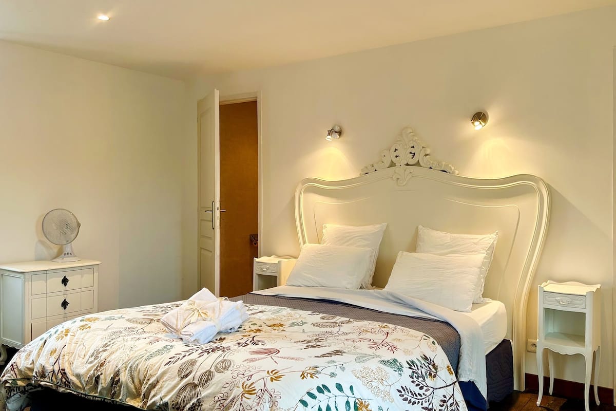 Un lit bien fait, recouvert d’une couvre-lit à motifs floraux, une table de chevet avec une lampe et une armoire avec un miroir.