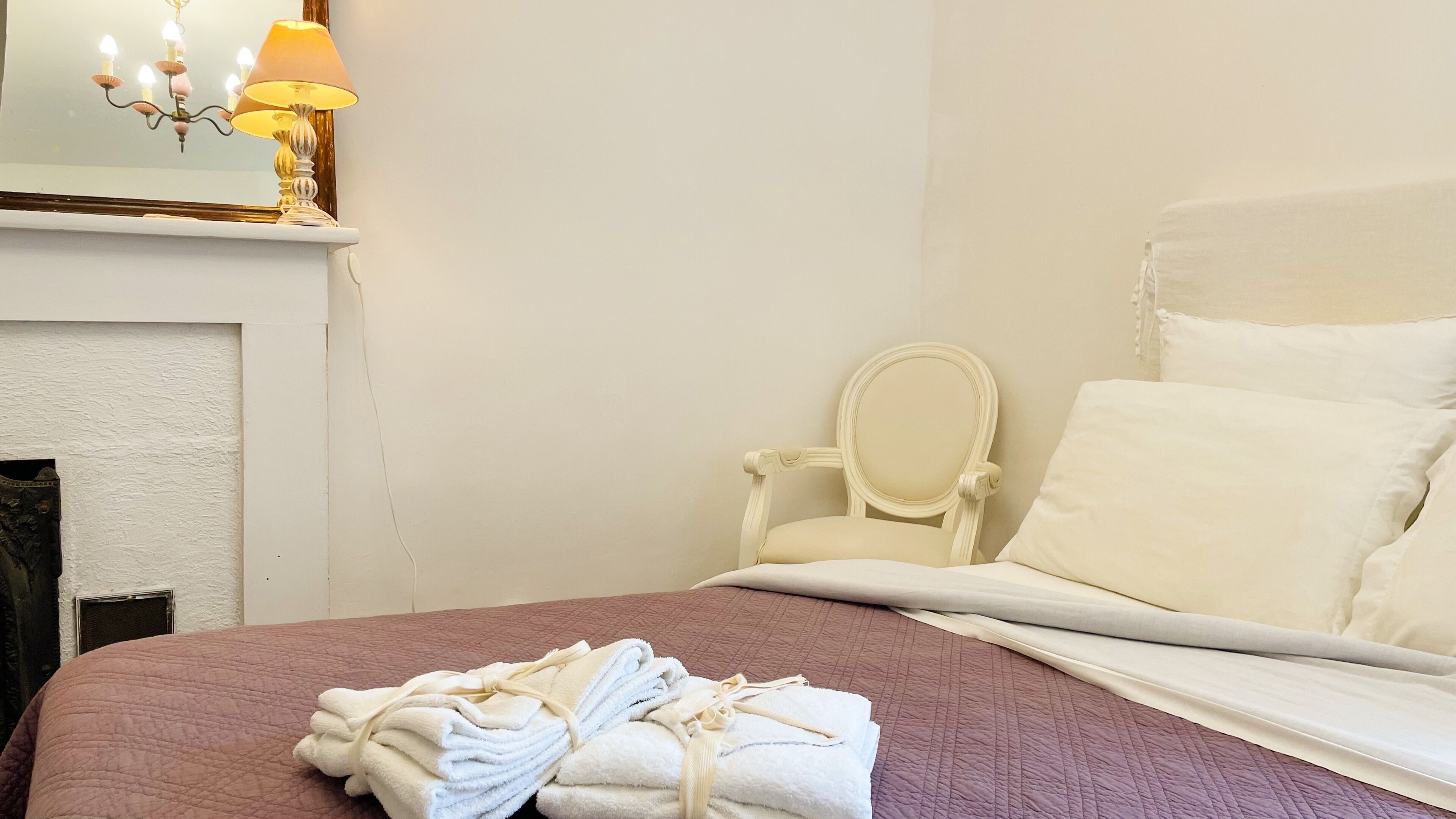 A neatly made bed with a pink quilt, a stack of white towels, and a white chair with a mirror.