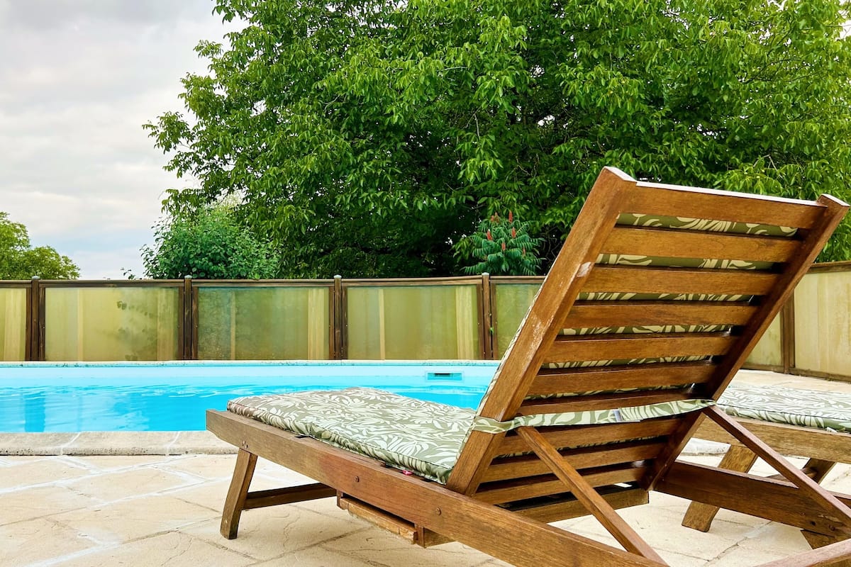 Un fauteuil de relaxation en bois avec un coussin, placé près d’une piscine, entouré d’une clôture et d’arbres.