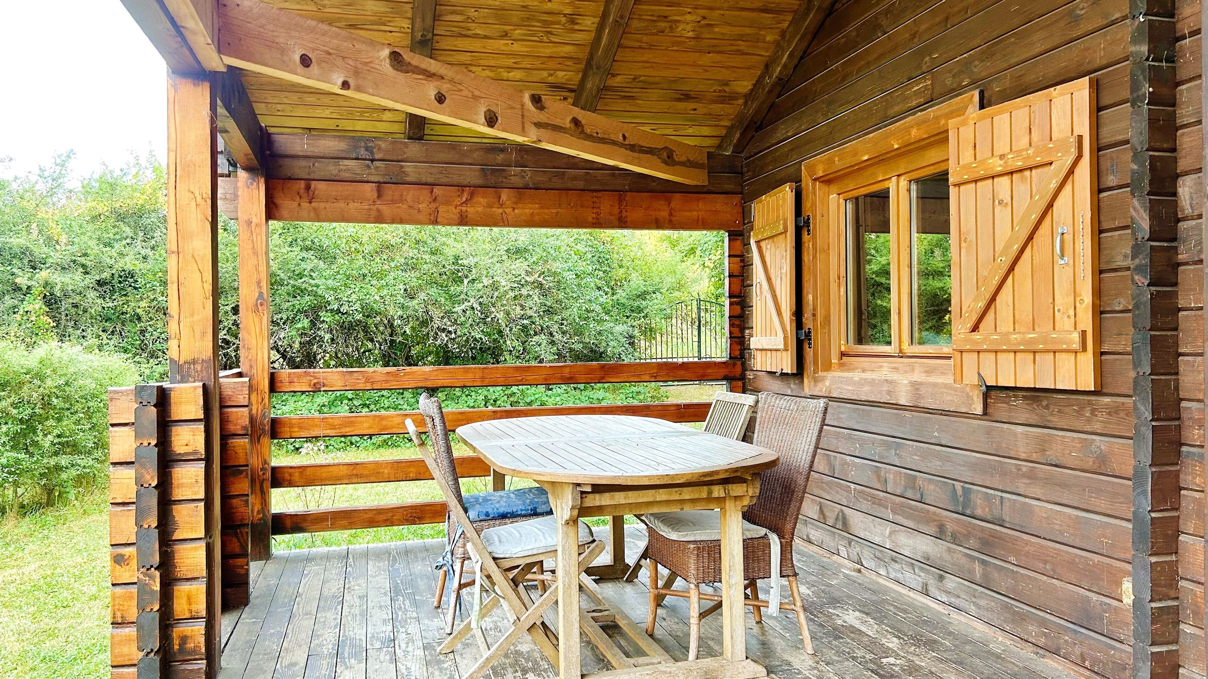 Eine Holzterrasse mit Tisch und Stühlen, ein Fenster mit Fensterläden und Blick ins Grüne.