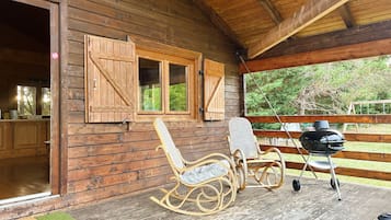 Eine Holzhütte mit Veranda, zwei Schaukelstühlen, einem Grill und einem Fenster mit Rollläden.