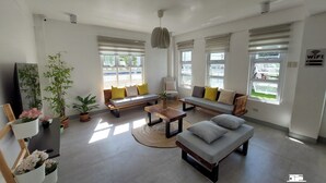 Un salon moderne avec un ensemble de canapés, une table basse et un téléviseur. On y trouve des plantes en pot et une grande fenêtre avec des stores.