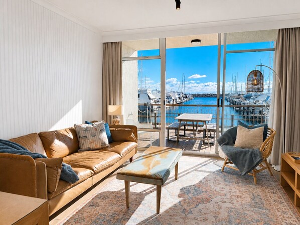 A modern living room with a leather sofa, a wooden coffee table, and a wicker chair. A large window offers a view of a marina with boats. - Amazing location on Fisherman’s Wharf only minutes to vibrant Fremantle nightlife and cafes. Enjoy the infamous West Australian Sunsets on your private balcony. (Fremantle)