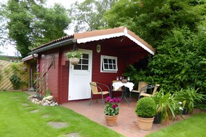 A red wooden house with a white door, a small porch with two chairs, and a table with a white tablecloth.