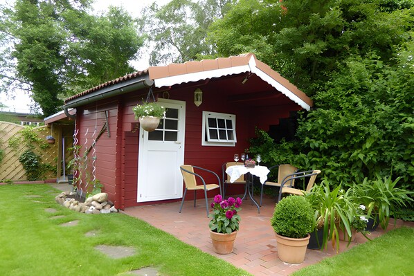 Ein rotes Holzhaus mit einer weißen Tür, einer kleinen Veranda mit zwei Stühlen und einem Tisch mit weißer Tischdecke.