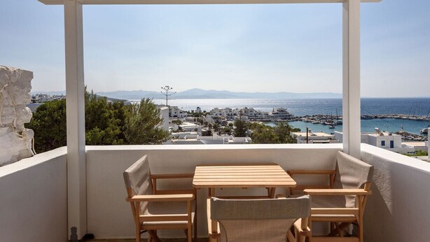 A balcony with a wooden table and chairs, overlooking a coastal town with buildings and a marina. - Phaedrus Living: Paros Cycladic Residences 6 (Piso Livadi)