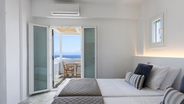 Une chambre d’hôtel avec un lit, une petite table et une vue sur la mer, visible par une porte ouverte.