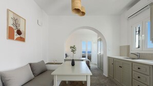 A modern kitchen with a central island, a dining area with a sofa, and a view of the sea through an archway. - Phaedrus Living: Paros Cycladic Residences 11 (Piso Livadi)