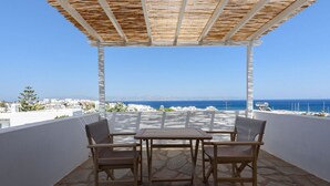 A covered outdoor seating area with a table and chairs, overlooking a coastal view with buildings and the sea. - Phaedrus Living: Paros Cycladic Residences 5 (Piso Livadi)