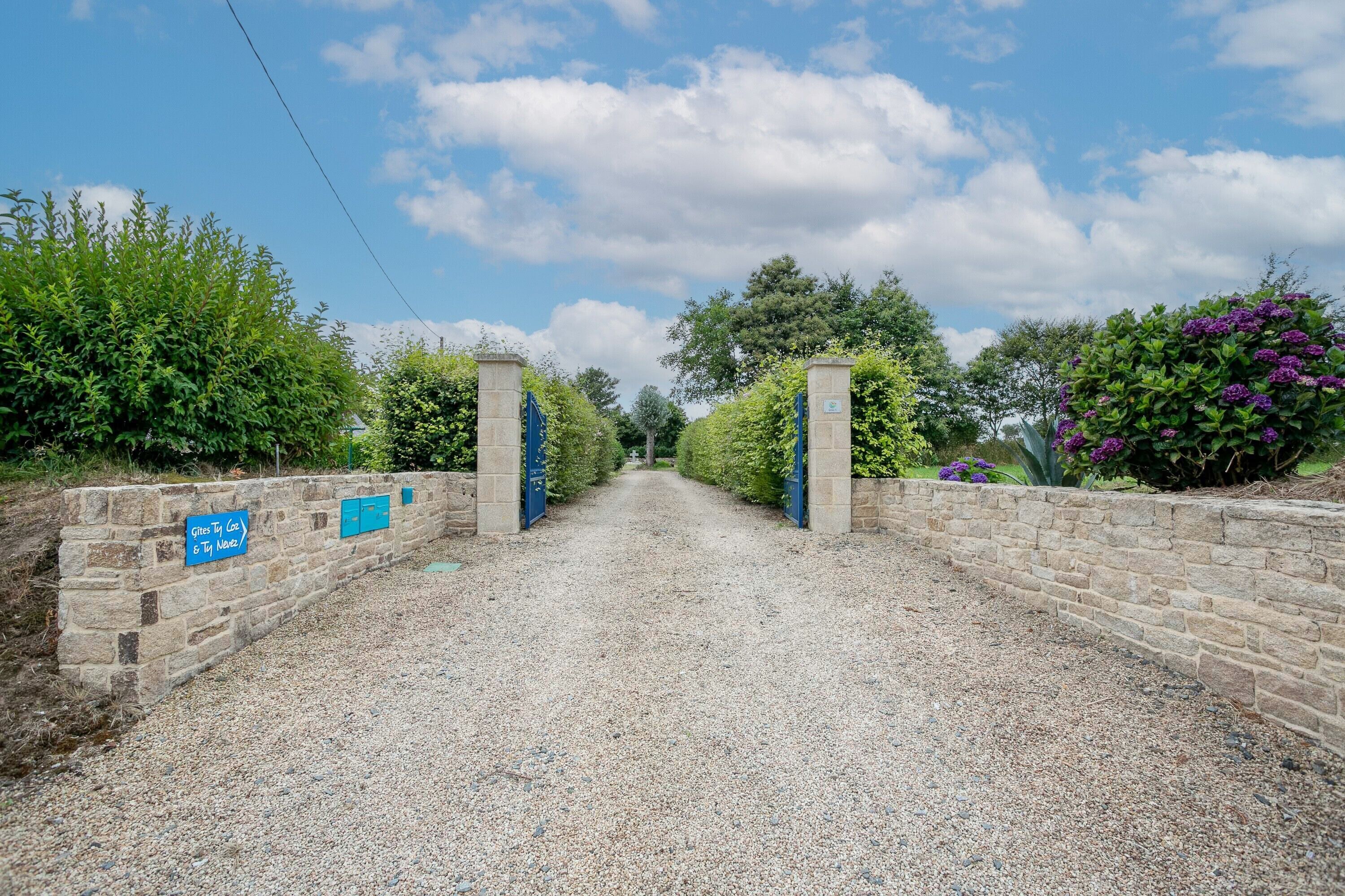 Un chemin de gravier mène à une entrée fermée par un portail, flanquée de murs en pierre et bordée de verdure de part et d’autre.