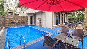 Una piscina con un paraguas rojo, sillas de mimbre y una mesa.