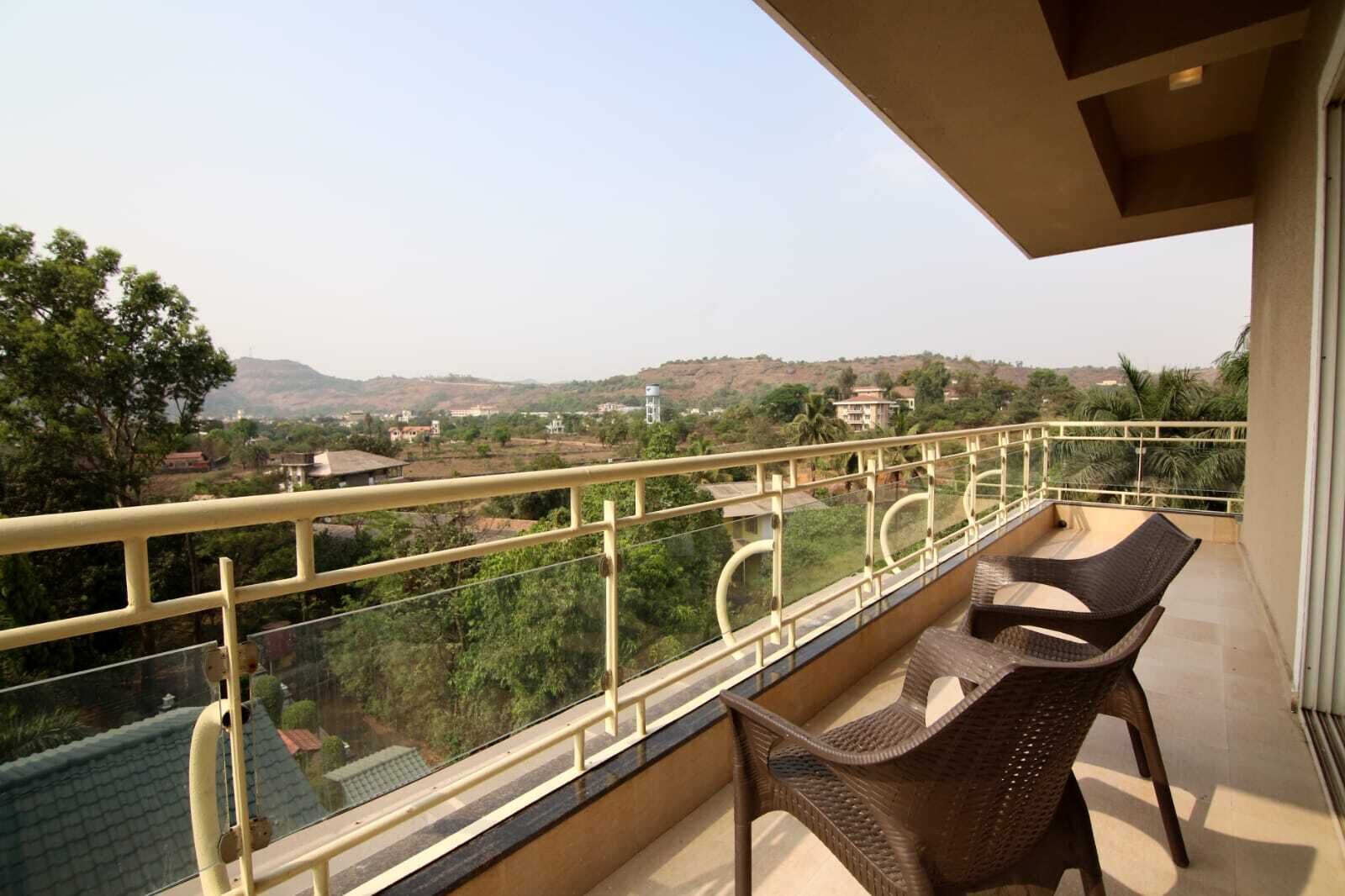 A balcony with two wicker chairs, a glass balustrade, and a view of a hilly landscape with buildings in the distance.