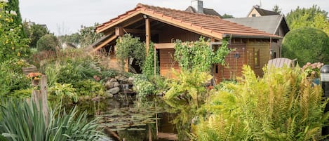 A wooden house with a tiled roof, surrounded by a garden with a pond and various plants.