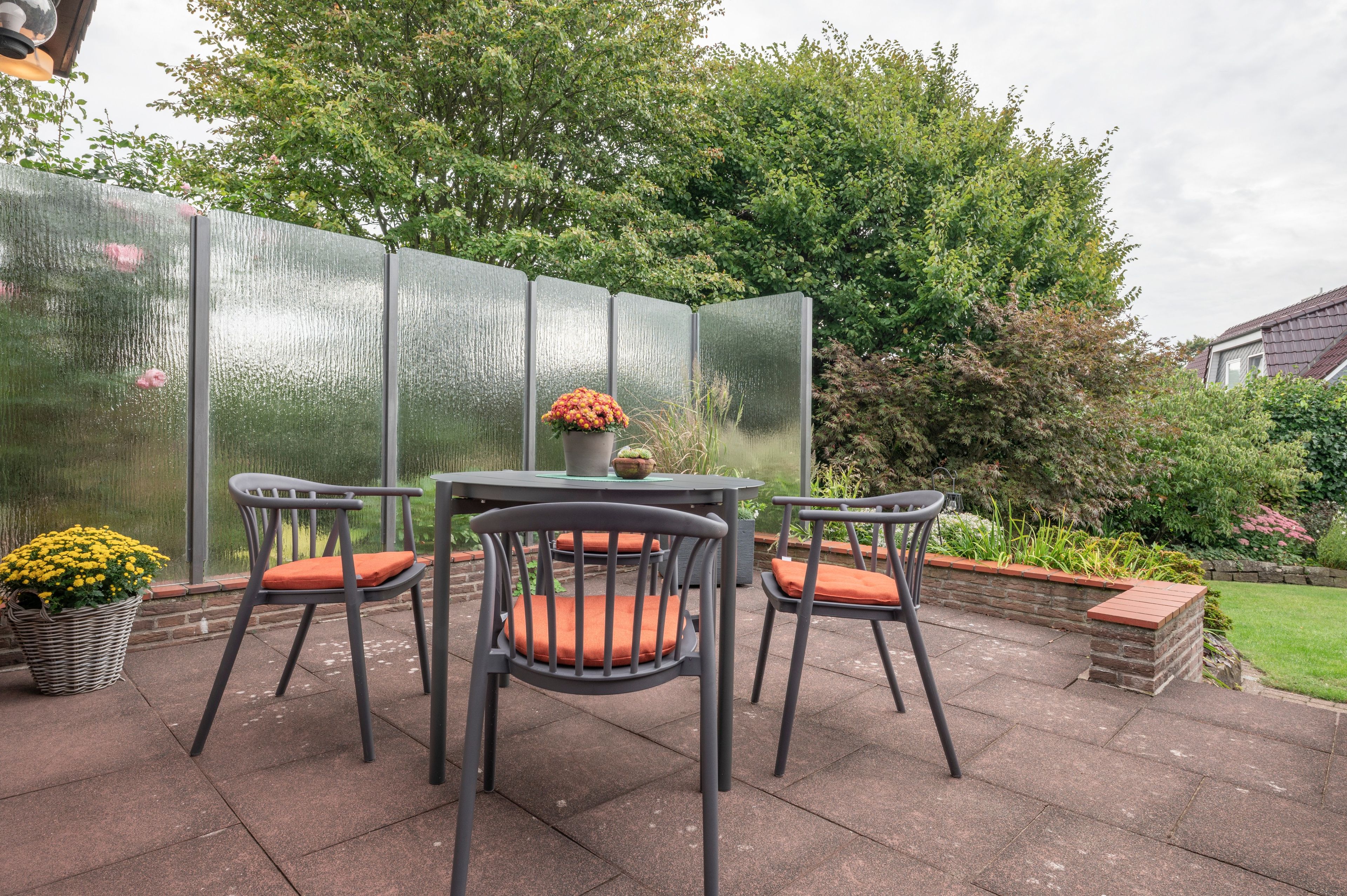 Eine Terrasse im Freien mit vier Stühlen und einem Tisch, umgeben von einer Glasscheibe und verschiedenen Pflanzen.