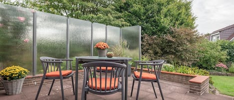 Eine Terrasse im Freien mit vier Stühlen und einem Tisch, umgeben von einer Glasscheibe und verschiedenen Pflanzen.
