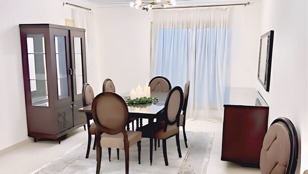 A modern dining room with a round table, six chairs, a chandelier, a cabinet, and a large window with curtains. - 2bedroom Apartment and Living Room at Shiekh Zayed (Sheikh Zayed City)