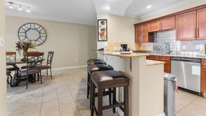 A kitchen with wooden cabinets, a stainless steel dishwasher, and a dining area with a table and chairs. - Two Bedroom with Pool Views (Destin)