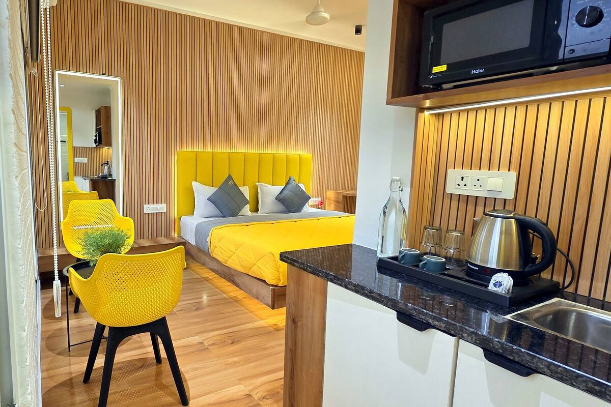A modern hotel room with a bed, a yellow chair, a small table, and a kitchenette.