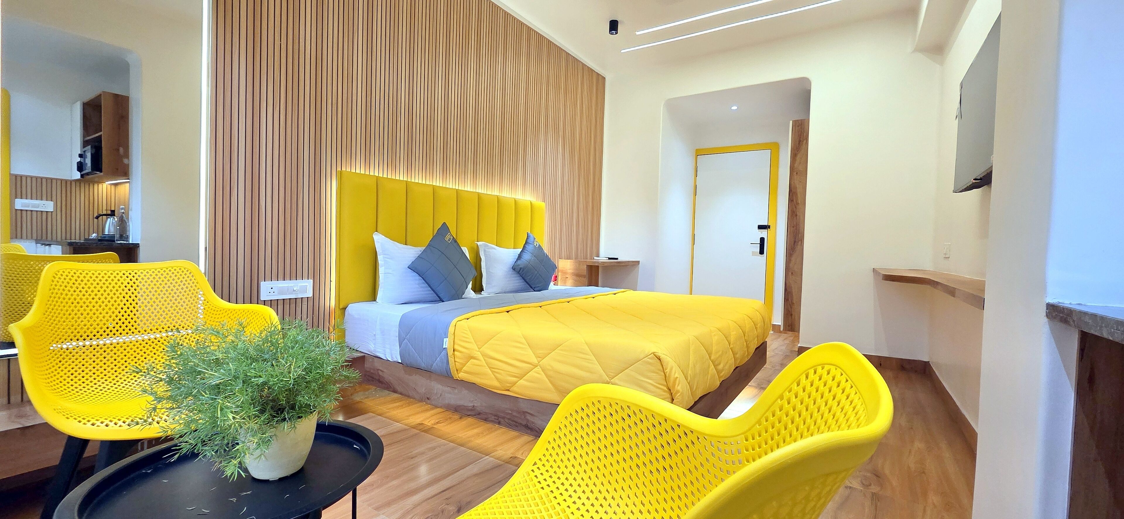 A modern hotel room with a bed, two yellow chairs, a small table with a plant, and a wooden accent wall.