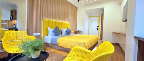 A modern hotel room with a bed, two yellow chairs, a small table with a plant, and a wooden accent wall.