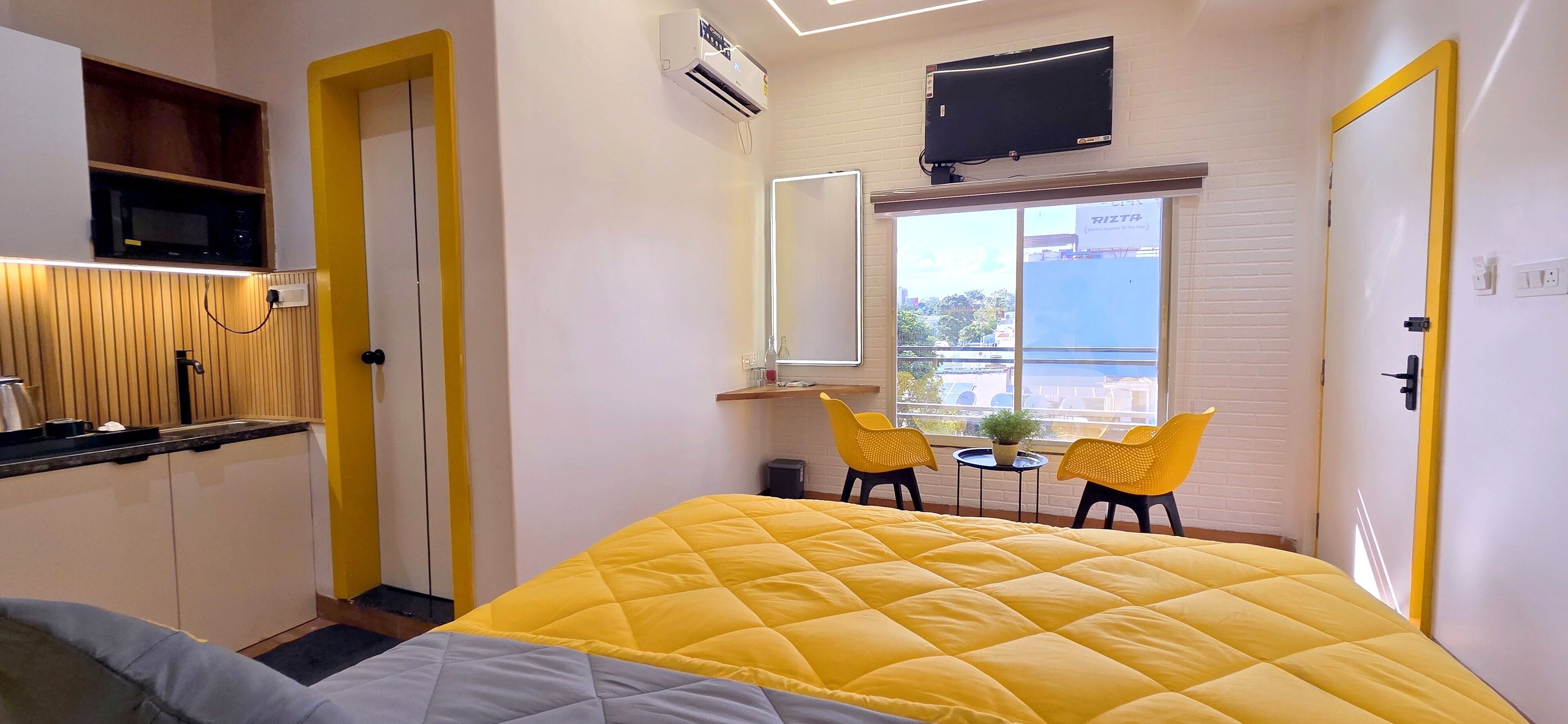 A compact hotel room with a yellow bed, a small kitchenette, a TV, a window with a view, and two chairs by the window.
