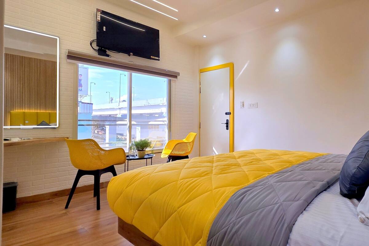 A modern hotel room with a large bed, a desk with a chair, a TV mounted on the wall, and a window with a view of the city.