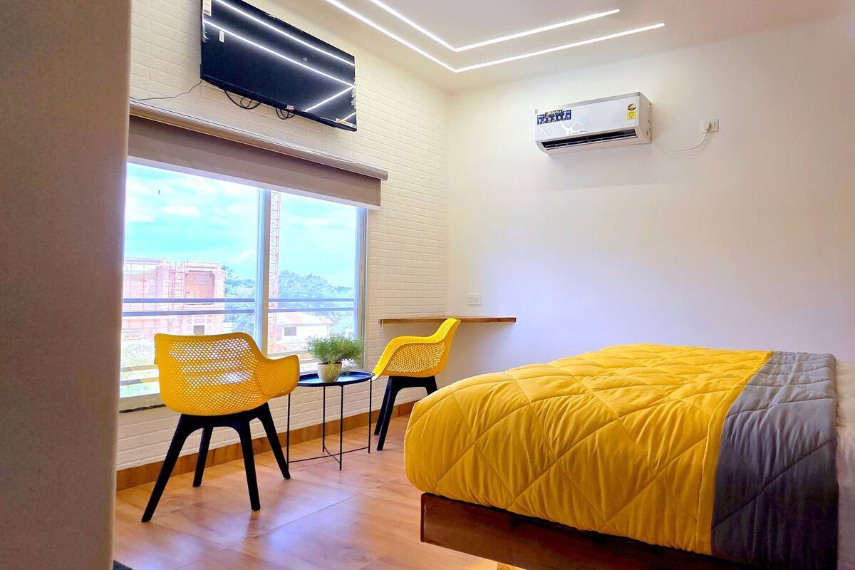 A modern hotel room with a large bed, a window with a view, a small table, two chairs, and an air conditioning unit.
