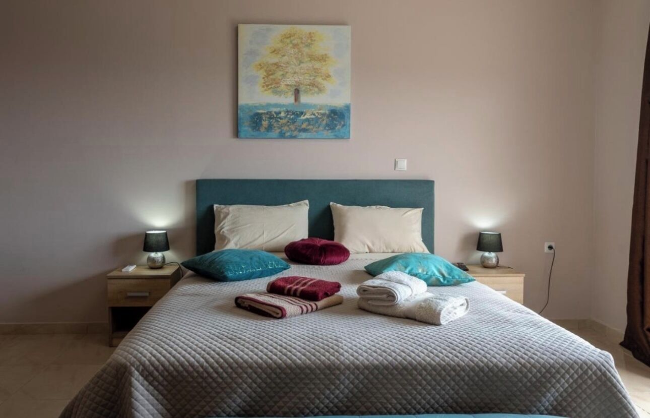 Een net opgemaakt bed met een gewatteerde deken, twee kussens en een gevouwen handdoek. Boven het bed hangt een schilderij van een boom.