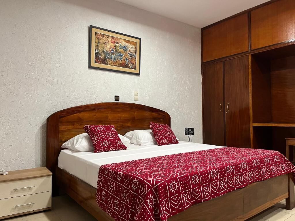 Ein Schlafzimmer mit hölzernem Kopfteil, einem Bett mit weißer und roter Bettwäsche, einem Nachttisch, einem Kleiderschrank und einem Gemälde an der Wand.