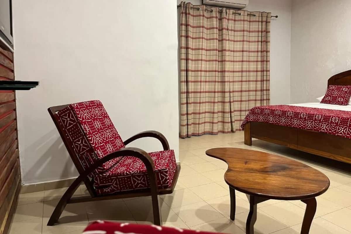 Una habitación pequeña y funcional con cama, una silla, una mesita y una cortina.