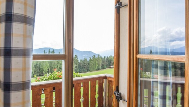 Une porte en bois ouverte donne sur un balcon doté d’une balustrade en bois et offrant une vue sur un paysage verdoyant, avec des montagnes à l’horizon.