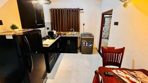 Une cuisine compacte équipée d’un réfrigérateur, d’une cuisinière et d’un évier. On y trouve une table à manger avec des chaises et une petite poubelle.