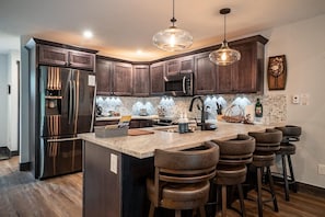 A modern kitchen with dark wood cabinets, stainless steel appliances, a large island with stools, and pendant lighting.