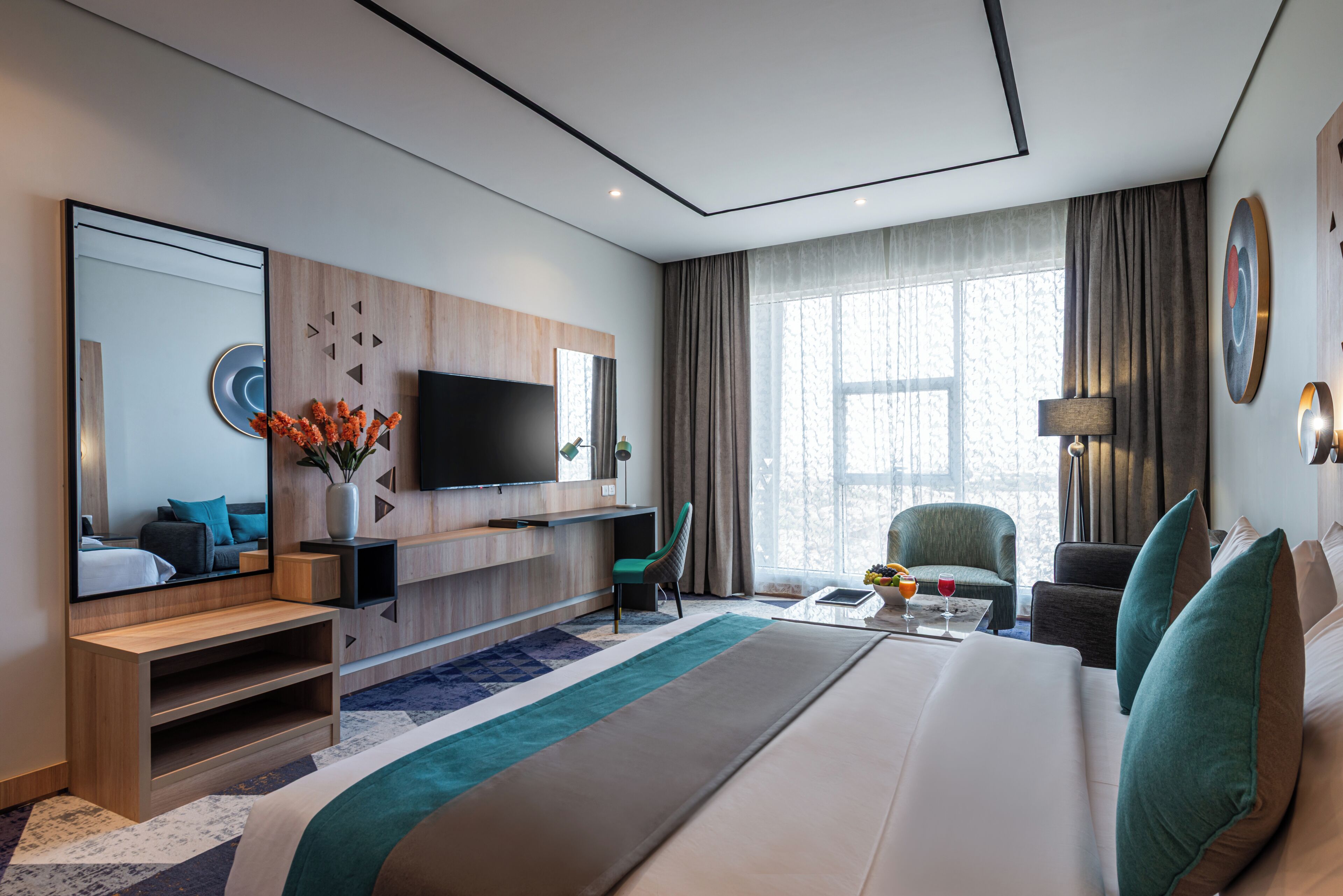 Una habitación de hotel moderna con una cama grande, un televisor de pantalla plana, una zona de estar con una mesa y un ventanal con cortinas.