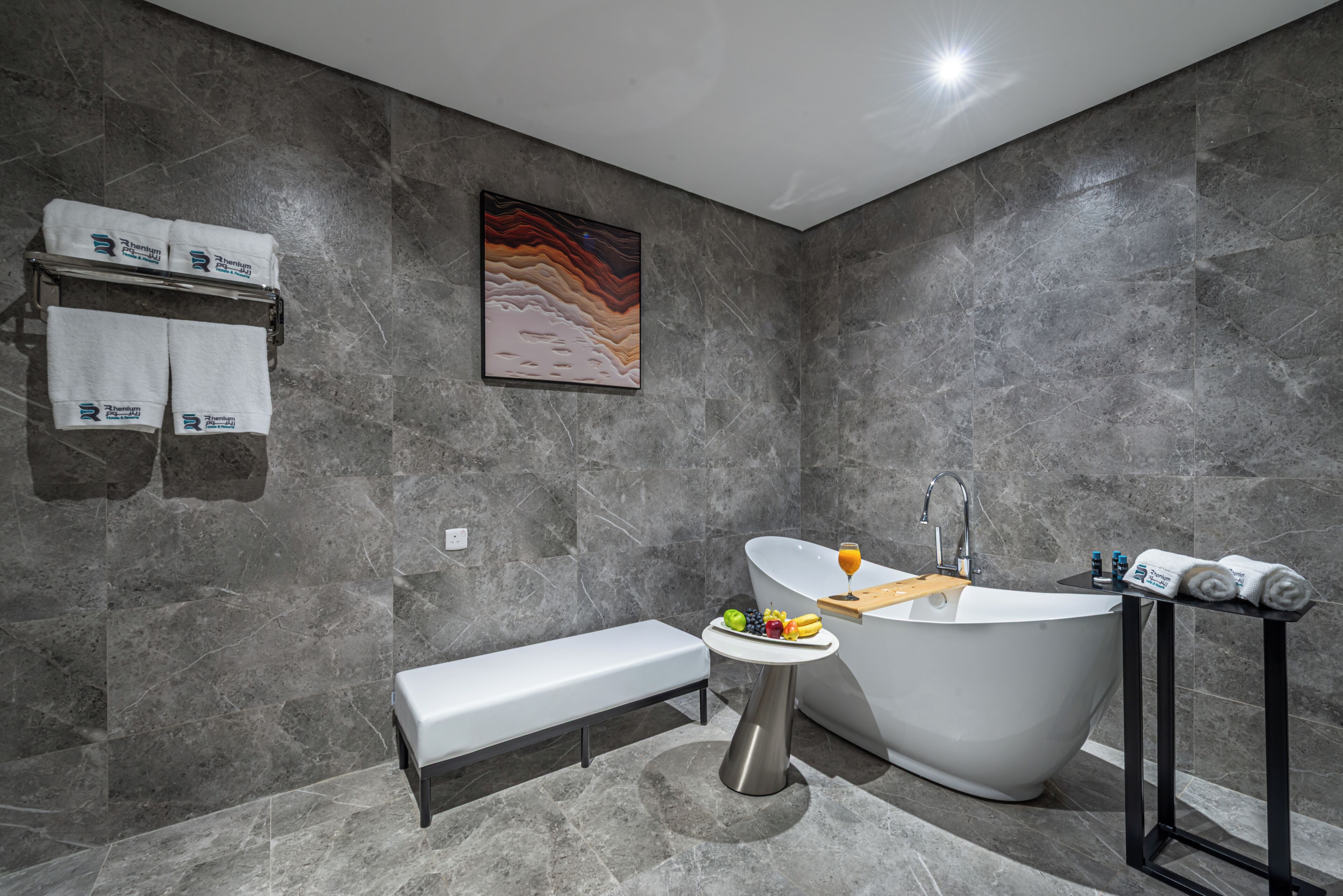 A modern bathroom with a freestanding bathtub, a bench, a small table with fruit, and towels on a rack.