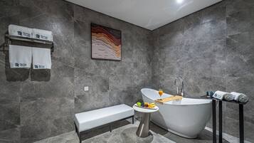 A modern bathroom with a freestanding bathtub, a bench, a small table with fruit, and towels on a rack.