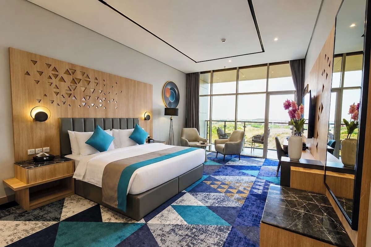 Una habitación de hotel moderna con una cama grande, un cabecero de madera, una alfombra estampada y vistas al exterior.