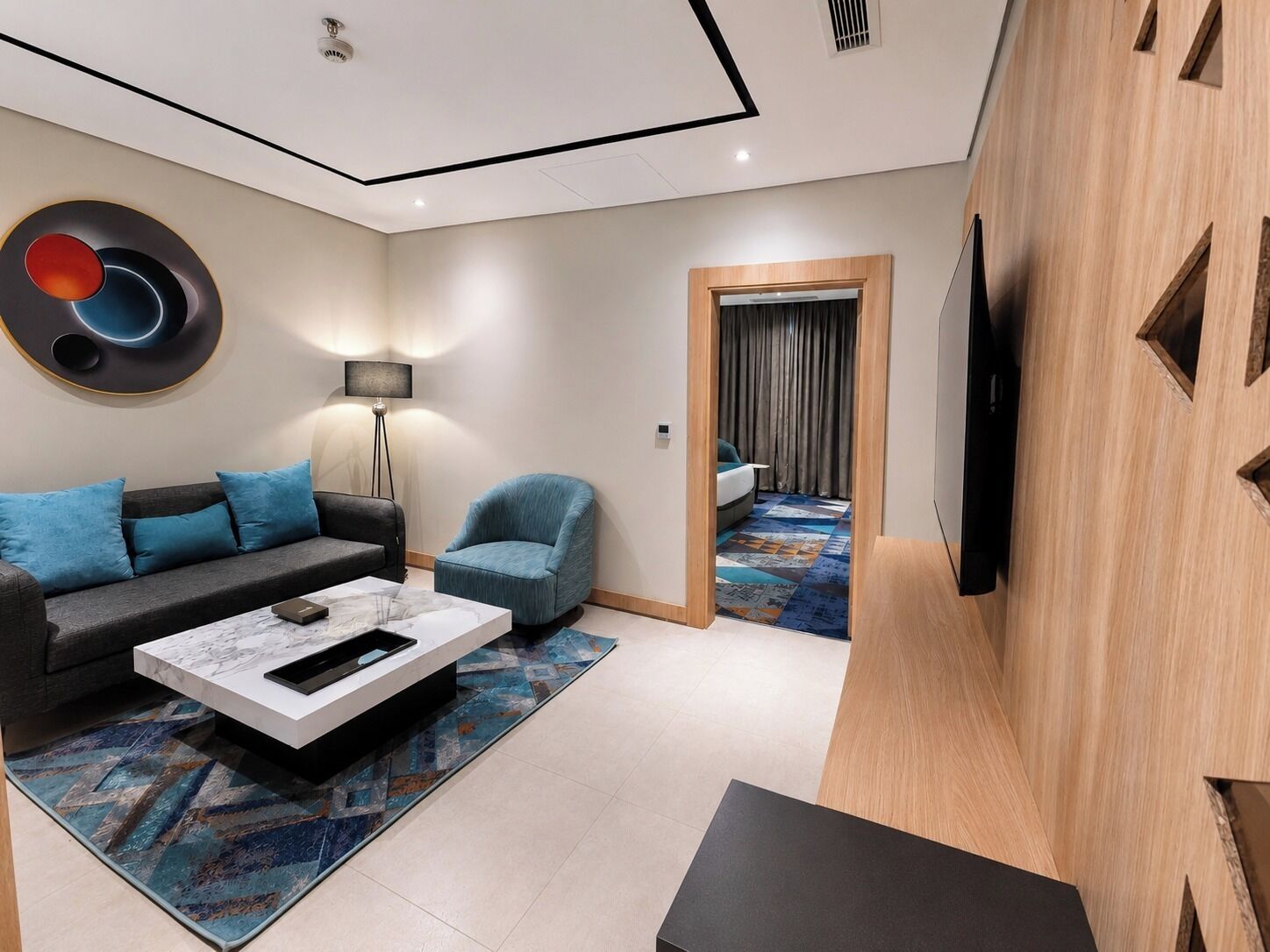 A modern hotel room with a sofa, armchair, flat-screen TV, and a patterned rug.