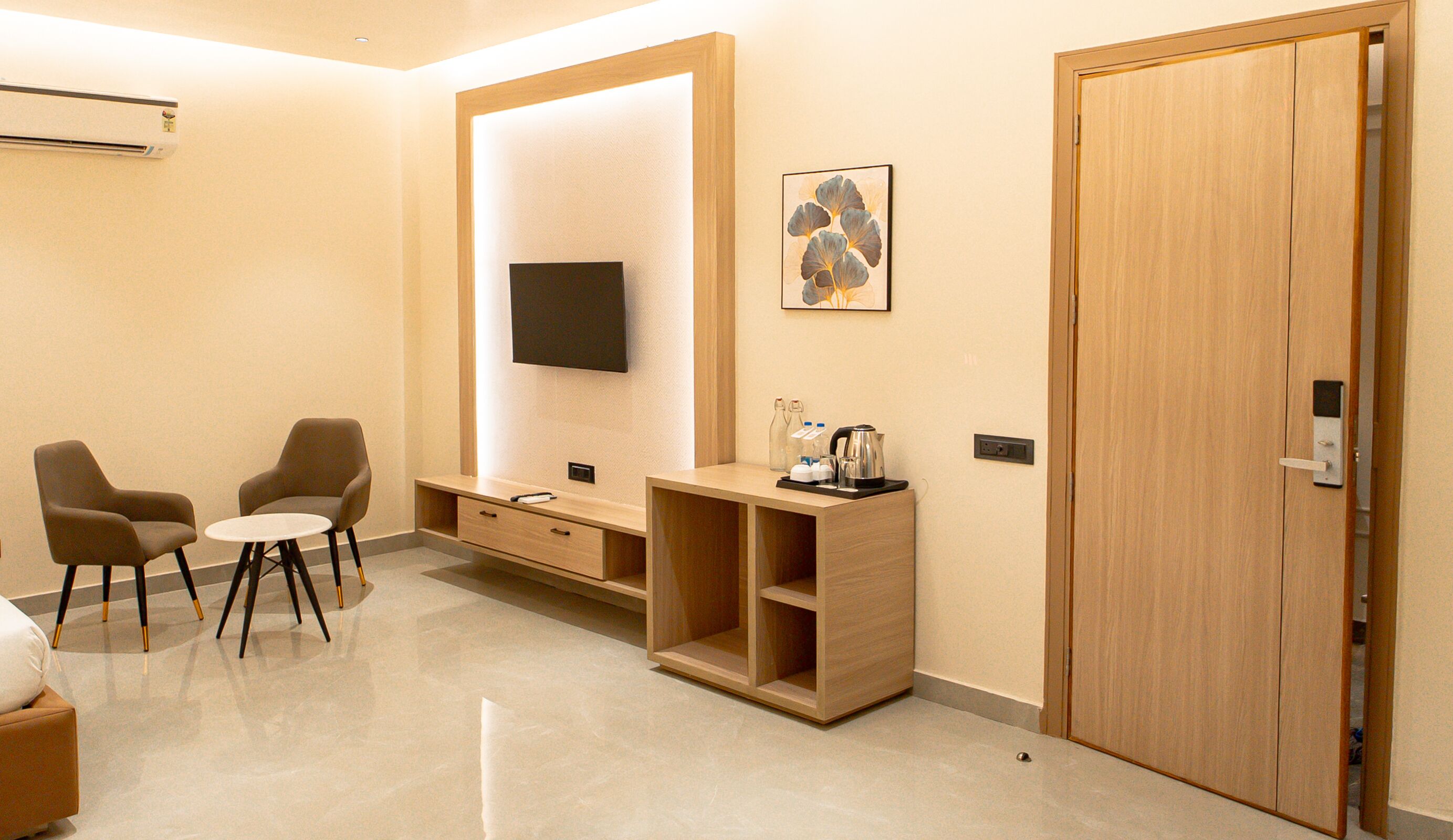 A modern hotel room with a flat-screen TV, a wooden desk with a coffee maker, two armchairs, and a large wardrobe.