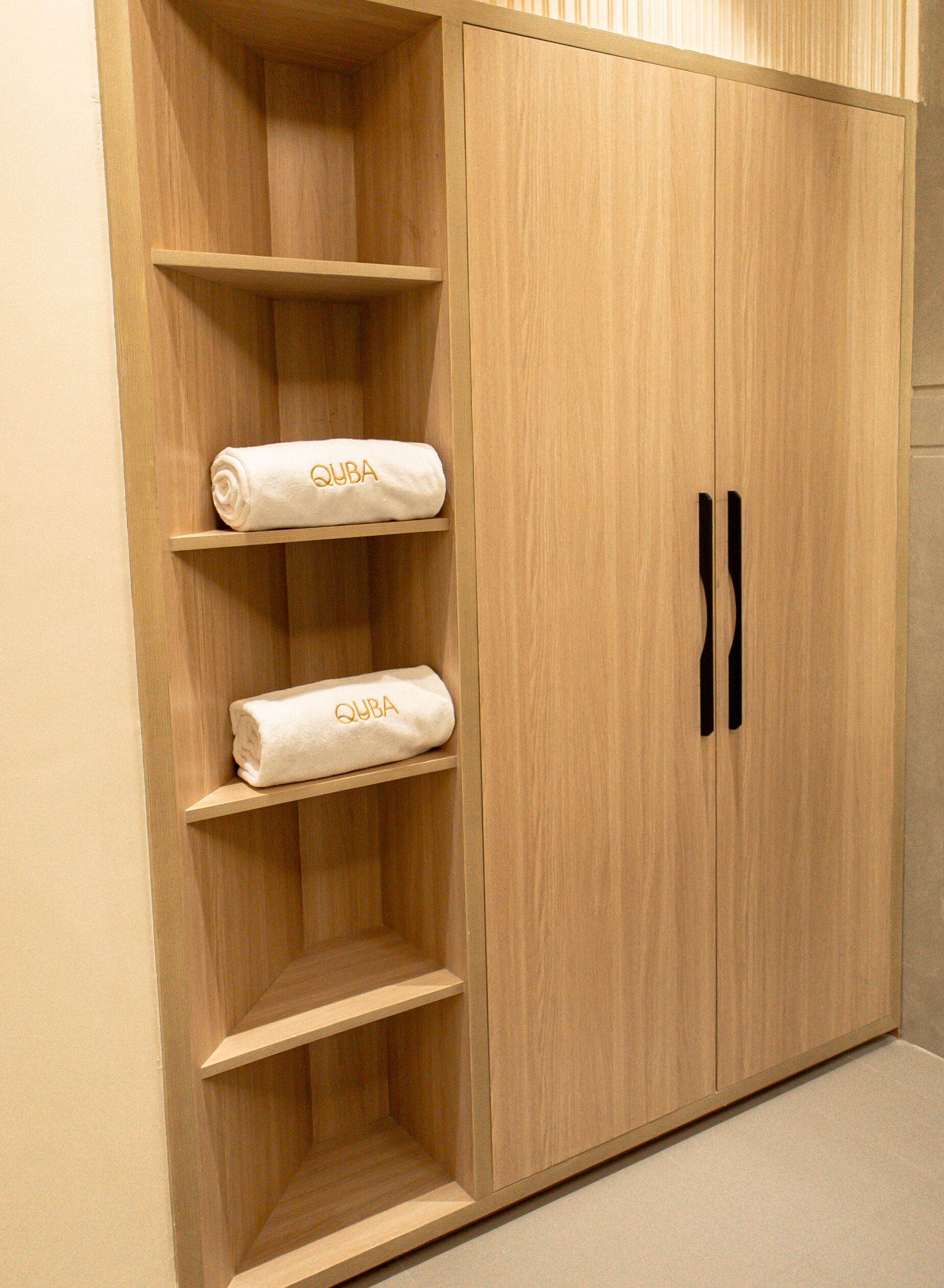 A wooden wardrobe with two doors and three shelves, each holding a rolled white towel with "QUBA" embroidered on it.