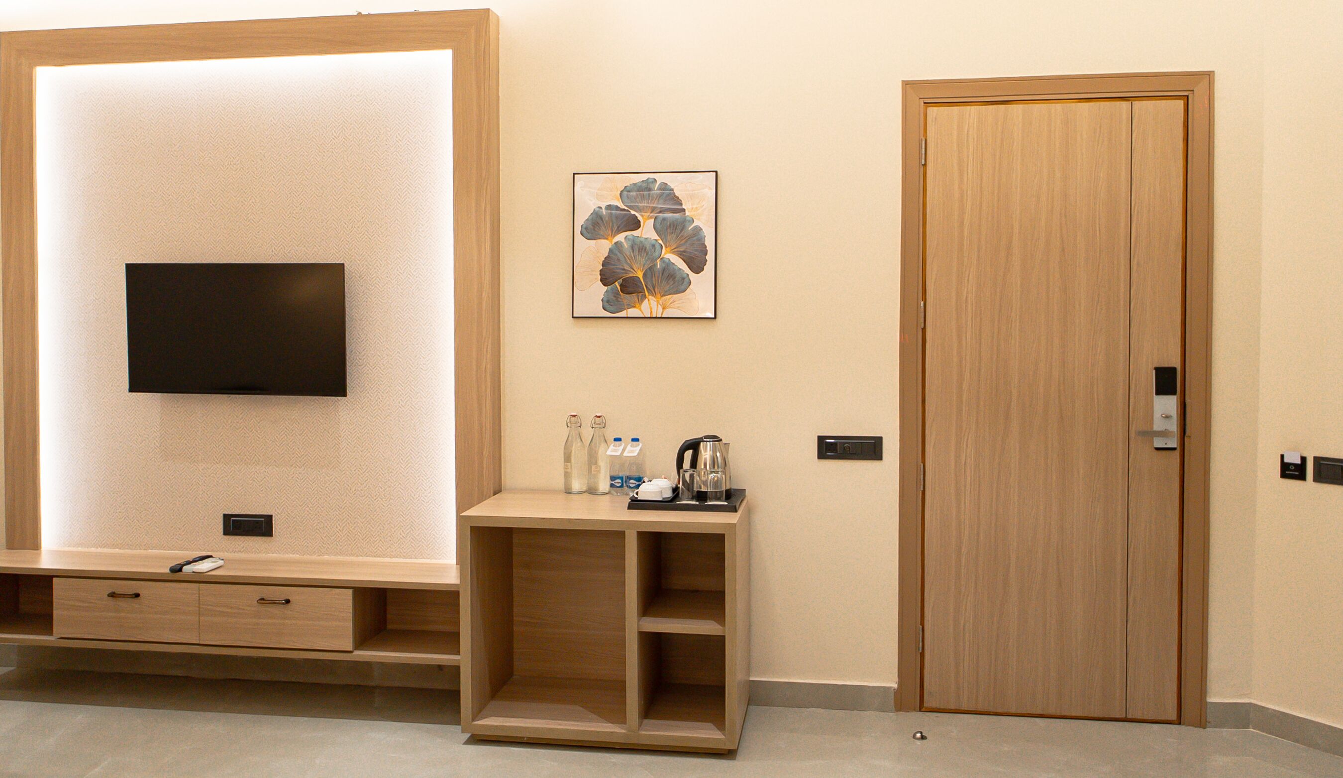 A hotel room with a flat-screen TV mounted on a textured wall, a wooden cabinet with a small table, a framed artwork, and a wooden door.