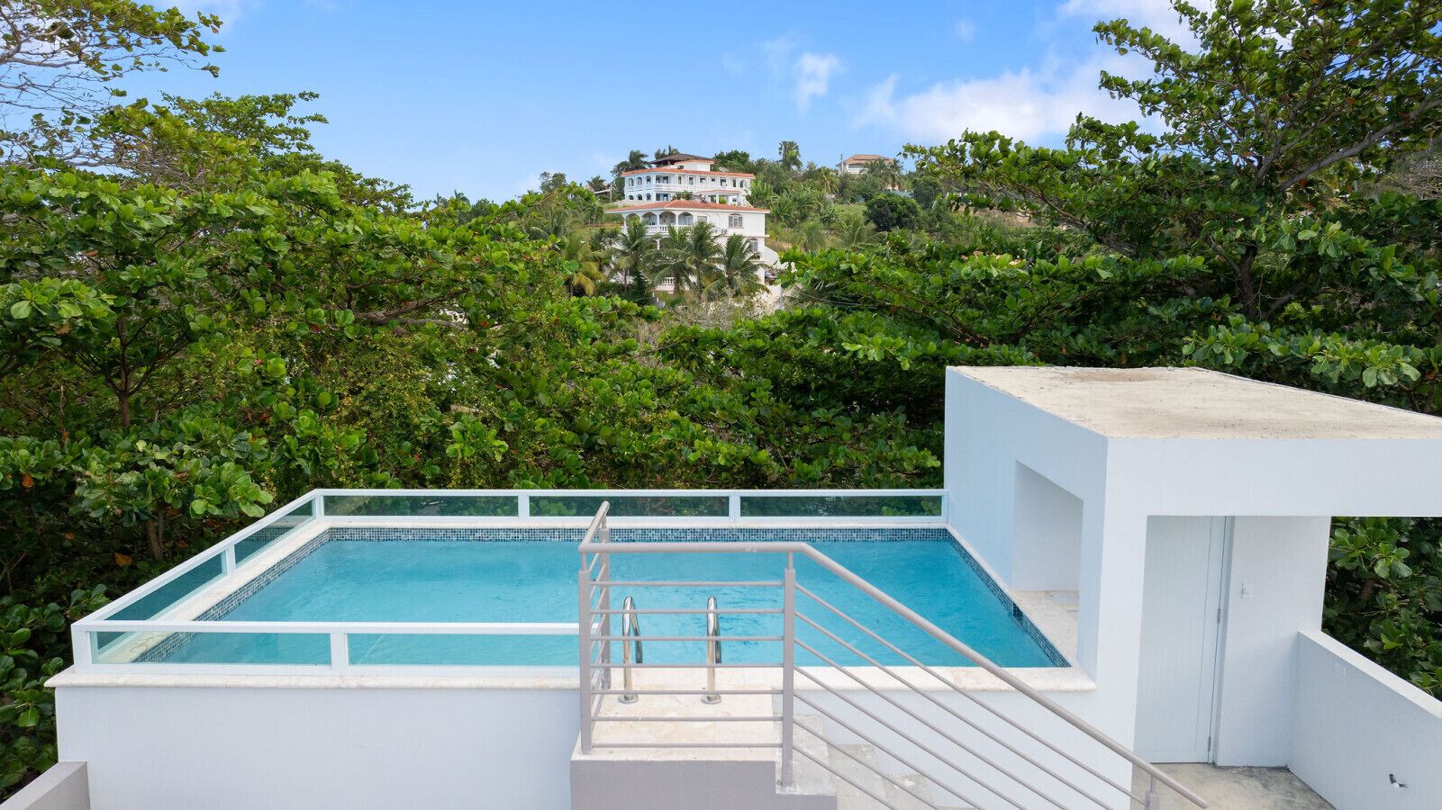 Piscina retangular com deck branco e grades de metal, cercada por vegetação exuberante e um edifício ao longe.