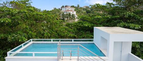 Piscina retangular com deck branco e grades de metal, cercada por vegetação exuberante e um edifício ao longe.