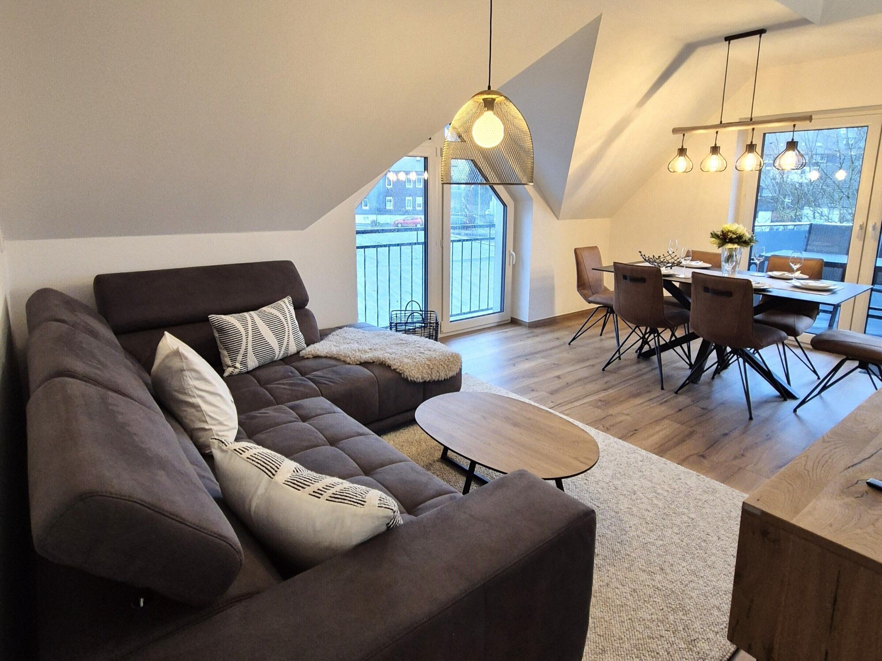 A modern living room with a sectional sofa, a dining area with a table and chairs, and a large window with a view of a balcony.